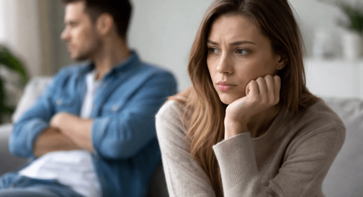 Woman looking sad and thoughtful while her ex boyfriend sits in the background appearing distant and angry after a breakup.