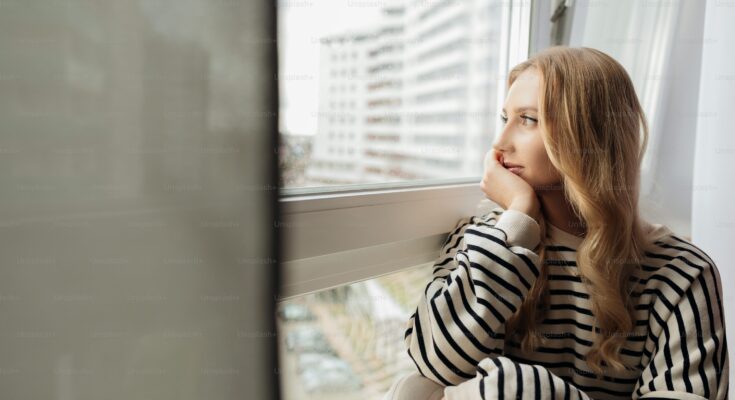 Woman looking thoughtfully out the window feeling distant from partner.