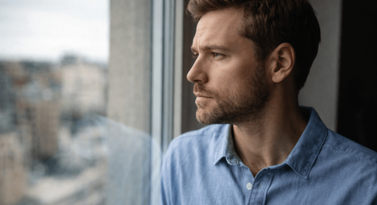 Man looking out a window with a thoughtful expression, representing the feeling of waiting after a breakup and wondering whether to reach out or stay silent.